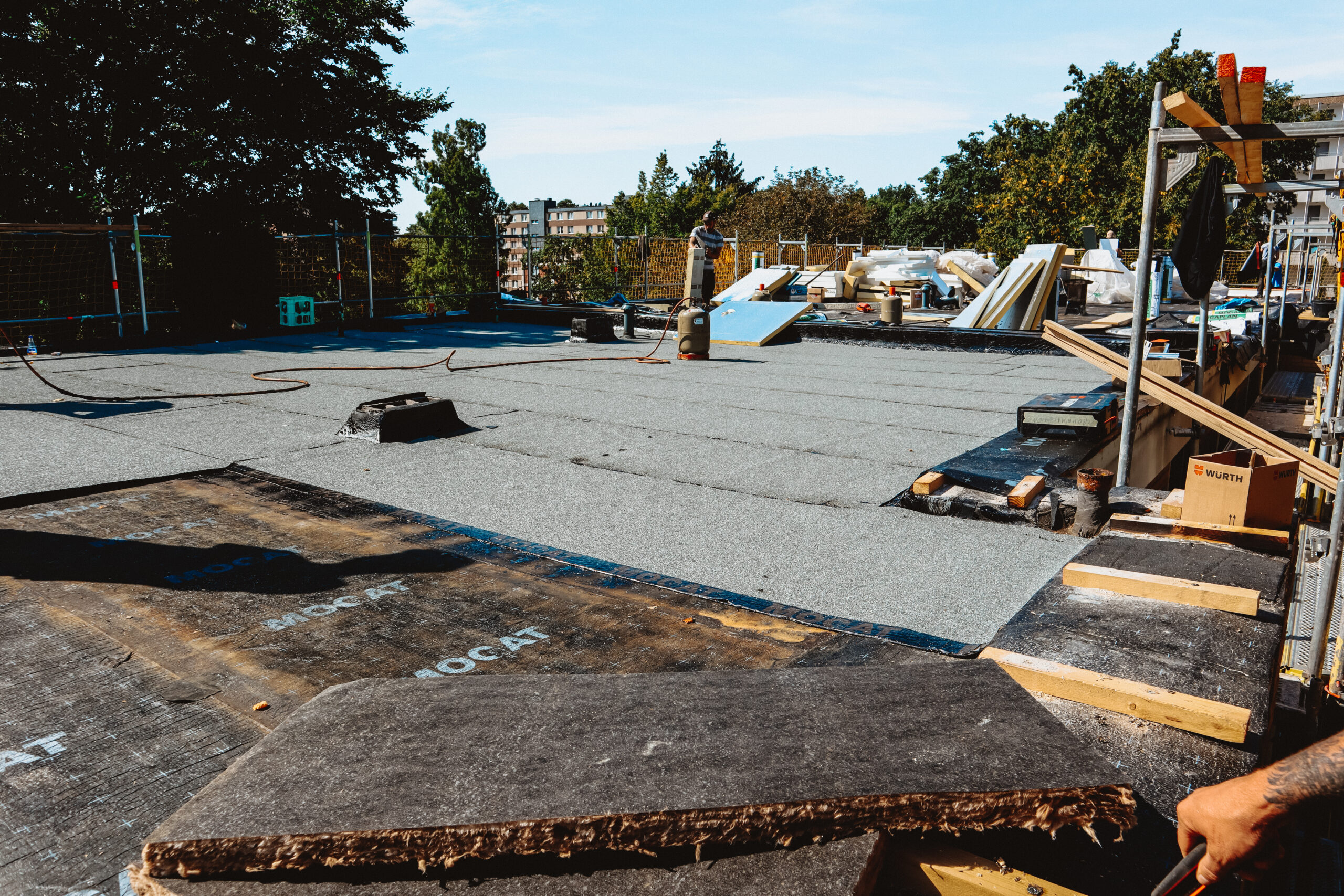 Das Bild zeigt im Rahmen der Bauleitung die Sanierung eines Flachdachs, wo gegenwärtig der Einbau von Dämmstoffeinlagen und die Applikation von Bitumenbahnen zu beobachten ist.